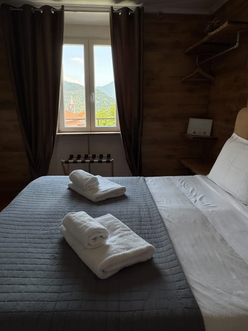 Camera da letto nella Collezione THE V, Sormano con vista montagna, asciugamani bianchi arrotolati sul letto, tende scure e paesaggio alpino