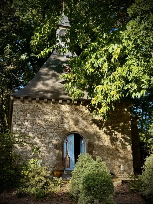 Chapelle en pierre avec porte bleue au Château de Montbrault, entourée d'arbres verts et de lierre, Fleurigné