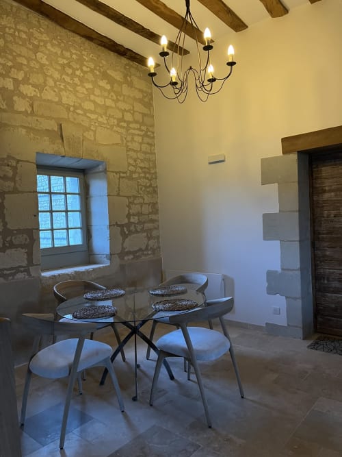 Salle à manger du gîte 1694 avec murs en pierre, poutres en bois, table ronde en verre, chaises modernes et lustre élégant