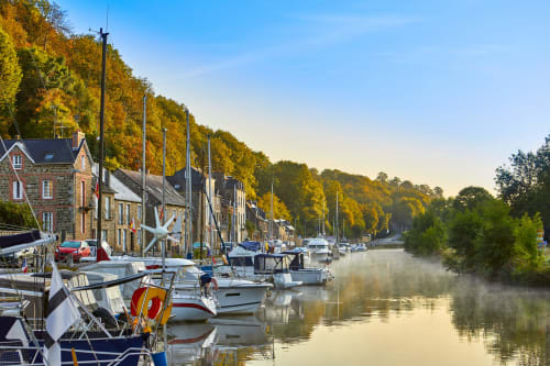 Voiliers amarrés le long de la rivière Arguenon au lever du soleil, bâtiments en pierre et arbres d'automne bordant le front de mer