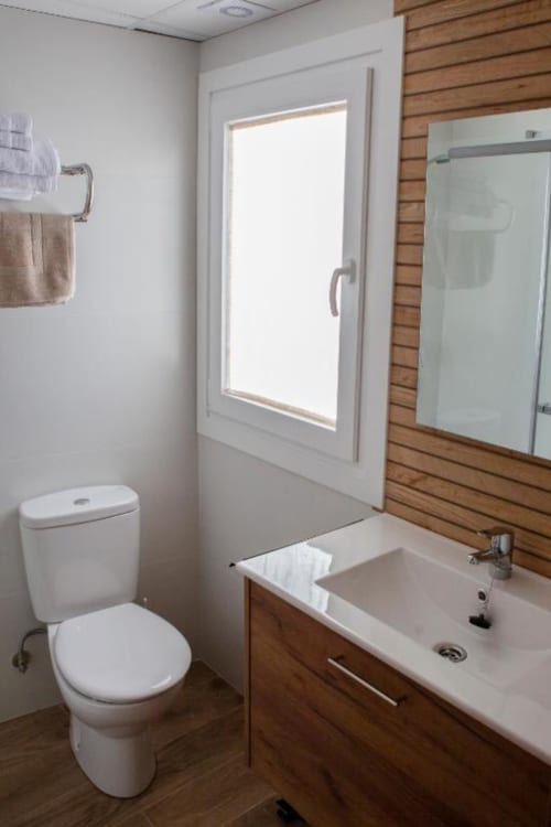 Baño moderno del Hostal Restaurante Angel con inodoro blanco, vanidor de madera, lavamanos, espejo y ventana luminosa