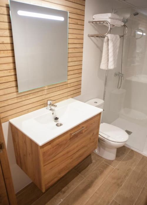 Baño moderno del Hostal Restaurante Angel en Huelma con tocador de madera, lavamanos blanco, espejo LED, inodoro y ducha