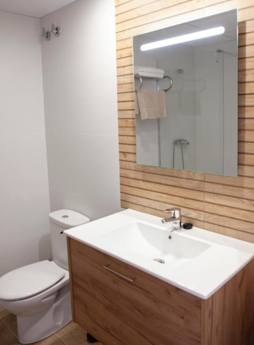 Baño moderno del Hostal Restaurante Angel con mueble de madera, lavabo blanco, espejo iluminado e inodoro en Huelma, Jaén