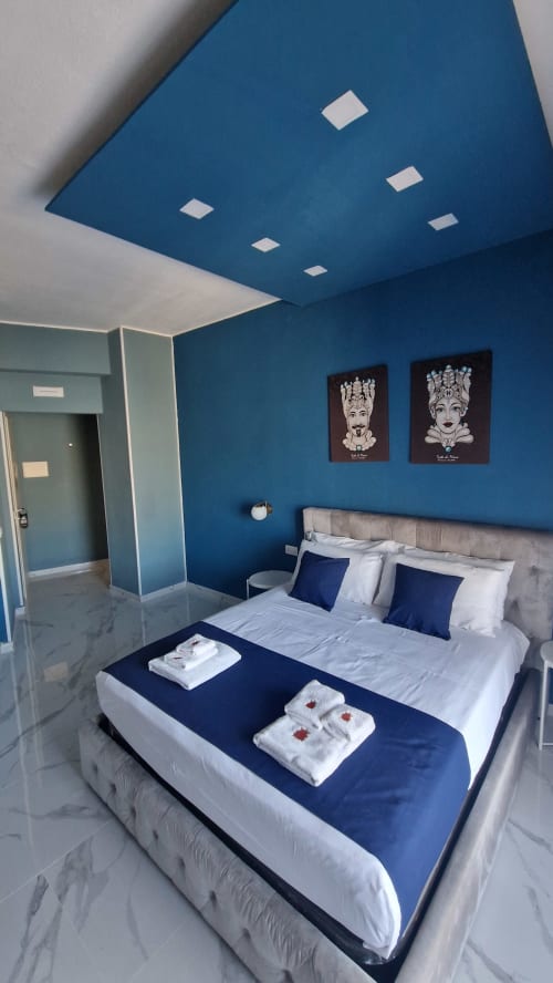 Camera da letto moderna e blu presso U Baruni B&B a Messina con letto bianco, cuscini scuri e arte contemporanea alle pareti