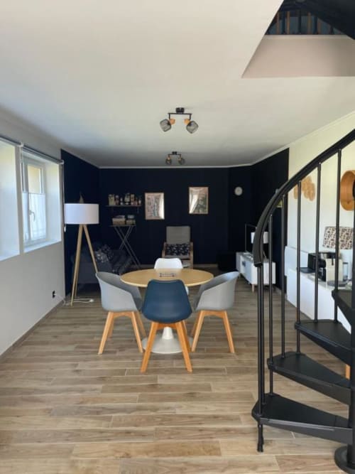 Salle à manger ouverte moderne du Gîte Le Clos d'Émilie avec table ronde en bois, chaises colorées, mur d'accent sombre et escalier en spirale