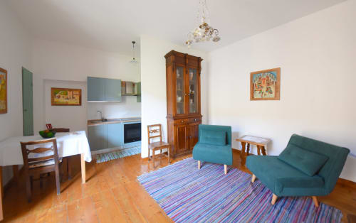 Panoramica dell'ampio soggiorno open space della Suite Drago al Roseto del Drago a Ponte in Valtellina (Sondrio): include poltrone relax, tipico pezzotto valtellinese, tavolo da pranzo, cucina a vista e quadri naïf dell'artista Marialuisa Cassan.