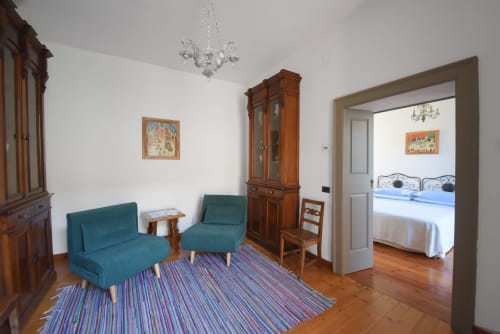 Vista del soggiorno con poltrone e pezzotto valtellinese affacciato sulla camera da letto matrimoniale della Suite Drago al Roseto del Drago a Ponte in Valtellina (Sondrio), impreziosita da mobili d'epoca e dipinti naïf di Marialuisa Cassan.