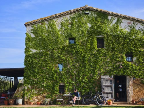 Antigua masía de piedra cubierta de hiedra en Llagostera con persona trabajando en mesa, bicicleta cercana, vistas a las montañas