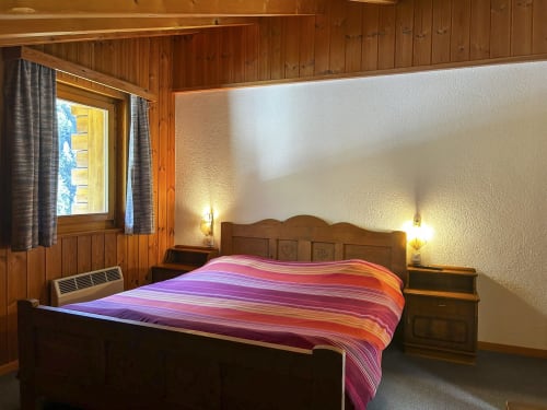 Chambre double cosy à l'hôtel Les Bouquetins à Zinal avec poutres en bois, literie rayée colorée et éclairage chaleureux
