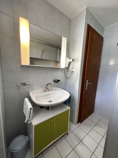 Salle de bains moderne à l'Hôtel les Bouquetins à Zinal avec carrelage blanc, meuble vert, miroir éclairé et porte en bois foncé