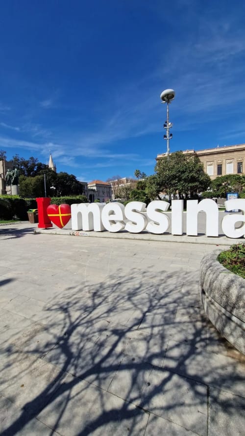 Cartello "I love Messina" in piazza pubblica circondata da edifici storici sotto un cielo azzurro a Messina, Italia