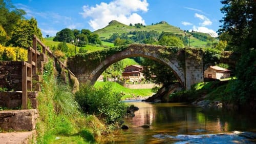 Puente de piedra con arco sobre río, colinas verdes y casas rurales del hotel La Calleja Real en Liérganes, España