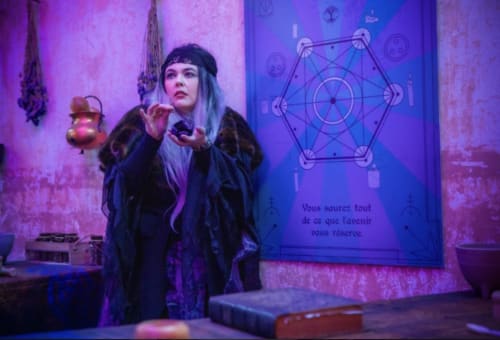 Figure mystérieuse en vêtements sombres tenant un objet orné contre un mur éclairé de violet avec symbole géométrique au Domaine de