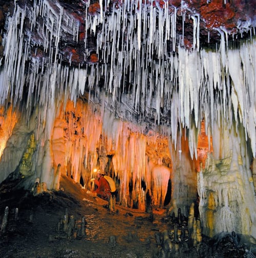 Cámara de cueva cubierta de carámbanos con depósitos minerales anaranjados y un espeleólogo explorando dentro del hotel La Calleja Real