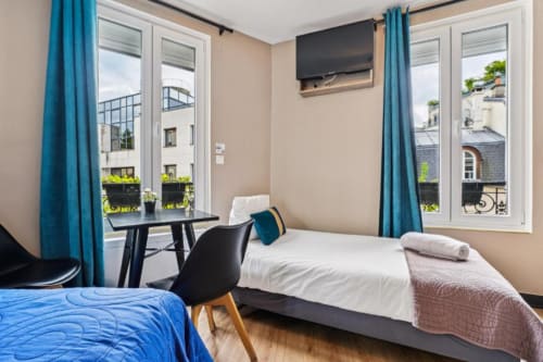 Bright studio bedroom at Résidence Aurmat Paris with twin beds, desk, balcony views, and blue curtains