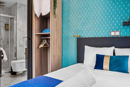 Modern studio bedroom at Résidence Aurmat Paris with turquoise patterned wall, white bed, blue pillows, and ensuite bathroom.