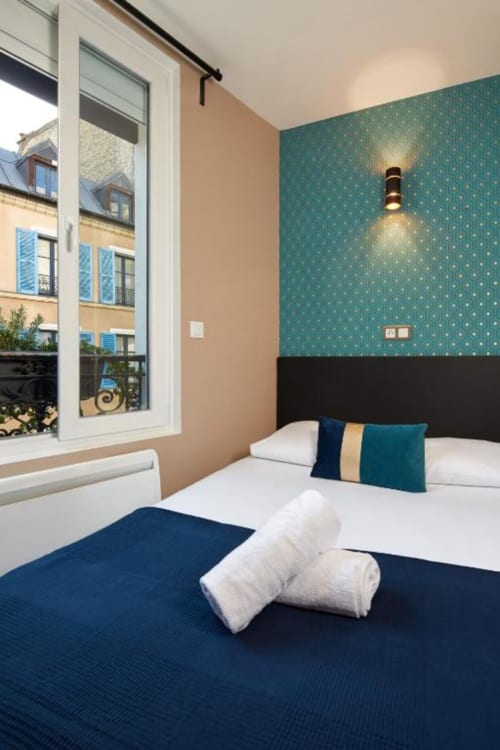 Chambre élégante de la Résidence Aurmat Paris avec papier peint à motifs bleus, literie marine et vue sur les rues parisiennes