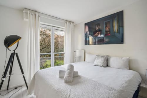 Chambre lumineuse d'un studio à la Résidence Aurmat Paris, vue sur jardin, literie blanche, lampadaire trépied, design moderne