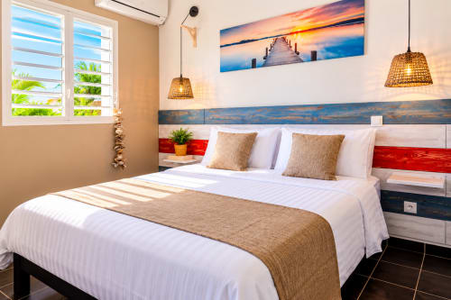 Chambre moderne au Quetzal à Sainte-Anne avec lit queen, luminaires suspendus tissés et tableau de ponton au coucher du soleil au-dessus de la tête de lit