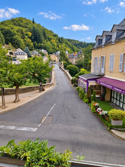 Vue pittoresque du village d'Estaing avec pont en pierre, bâtisse jaune et colline boisée, Hôtel du pont d'Estaing