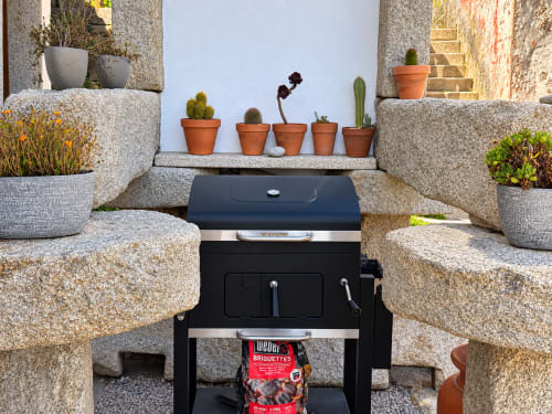 Parrilla Weber negra rodeada de asientos de piedra y cactus en macetas en jardín de cabaña costera en Holibai, As Mariñas