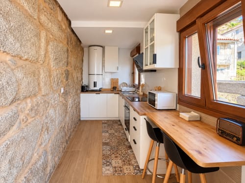 Cocina moderna en casita As Mariñas con muros de piedra, encimeras de madera, armarios blancos y escritorio junto a ventanas