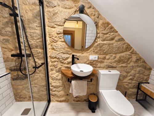 Baño moderno con muros de piedra rústica, espejo redondo, lavamanos blanco, inodoro y ducha walk-in en la casa rural Holibai en As