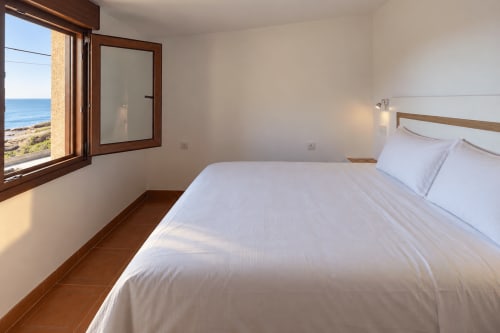 Luminoso dormitorio costero en la cabaña Holibai con vistas al mar, ropa de cama blanca, detalles en madera y piso de terracota