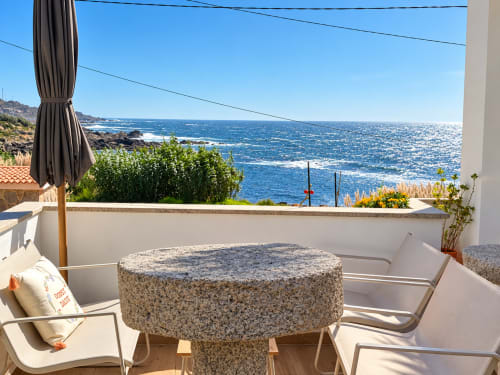 Terraza de casa costera con vistas al mar, mesa de piedra, tumbonas y sombrilla frente a aguas azules en As Mariñas, España