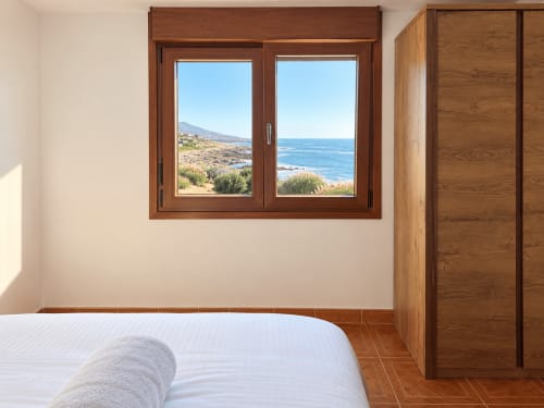 Dormitorio de cabaña costera en As Mariñas con vistas al mar por ventana de madera, cama blanca y armario de madera