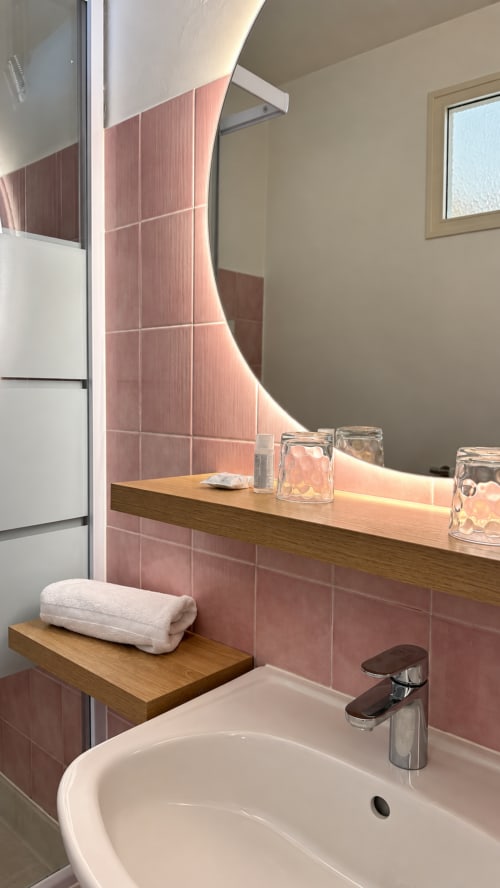 Salle de bains moderne de l'Hôtel Thalassa à Cargèse avec carrelage rose, meuble vasque en bois et miroir incurvé éclairé