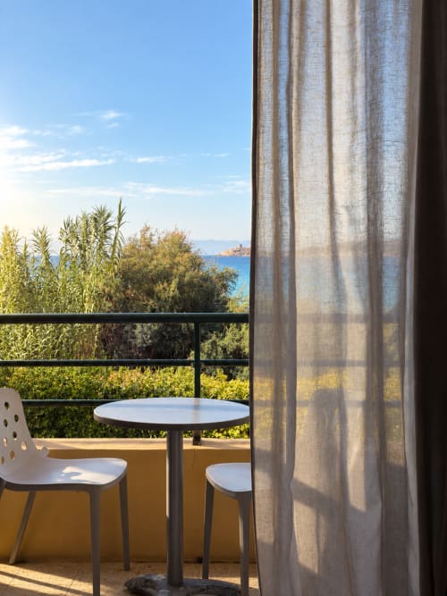 Vue du balcon de l'Hôtel Thalassa à Cargèse avec panorama marin, table moderne et chaises, rideaux fluides