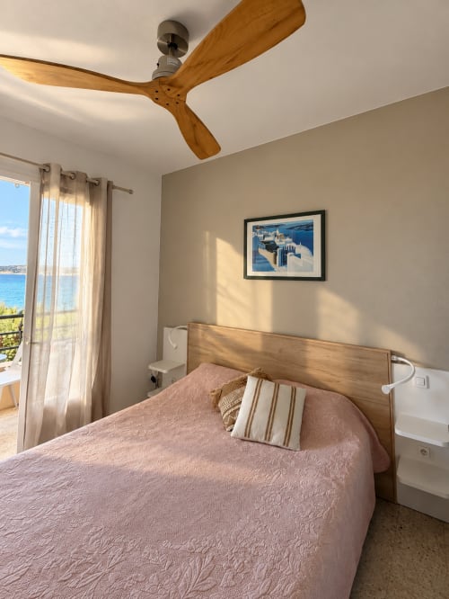 Chambre lumineuse à l'Hôtel Thalassa avec vue mer, ventilateur de plafond en bois et tableaux encadrés surplombant la côte de Cargèse