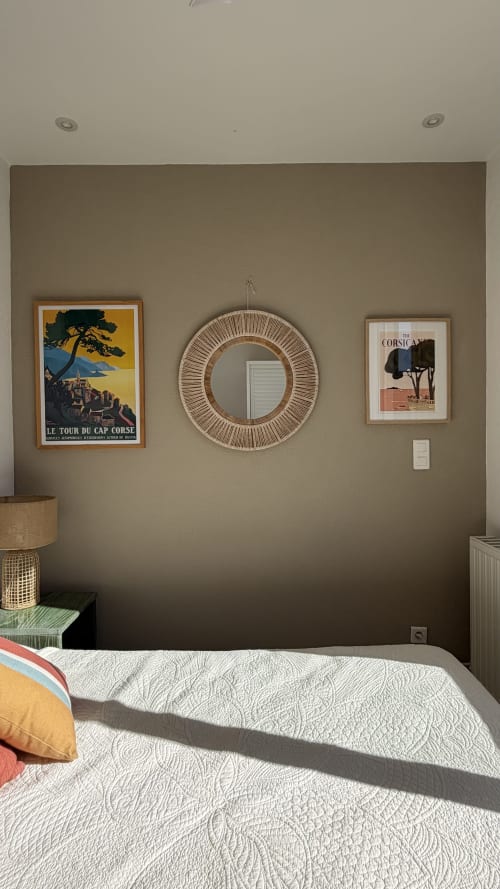 Chambre de l'Hôtel Thalassa à Cargèse avec murs beige, affiches vintage de Corse, miroir rond tressé et courtepointe blanche