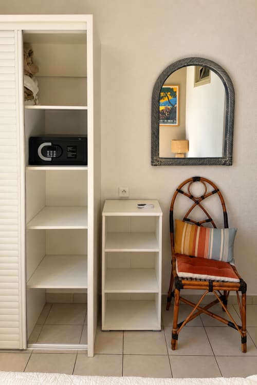 Détail de chambre Hôtel Thalassa : étagères blanches avec coffre-fort, petite table, miroir bombé et fauteuil en rotin avec coussins