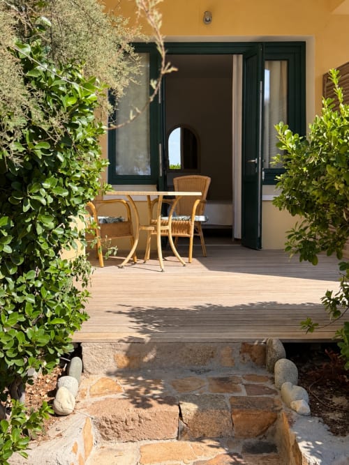Terrasse de l'Hôtel Thalassa à Cargèse avec terrasse en bois, chaises en rotin, plantes vertes et portes vitrées à cadres noirs
