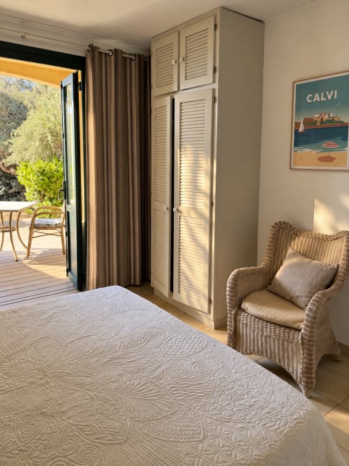 Chambre lumineuse à l'Hôtel Thalassa à Cargèse avec lit, fauteuil en osier, armoire persiennée et accès à la terrasse