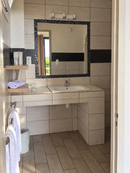 Salle de bains moderne de l'Hôtel Thalassa à Cargèse avec carrelage beige, miroir encadré de mosaïque noire, lavabo et étagères en bois