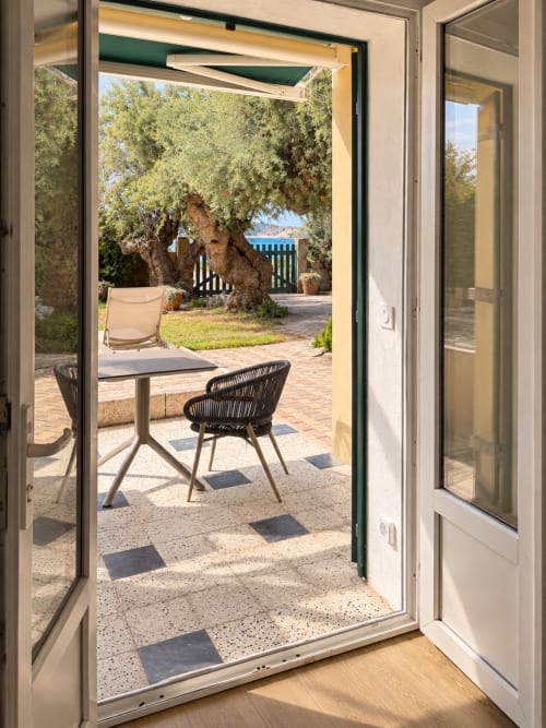 Terrasse de l'Hôtel Thalassa Cargèse vue par les portes vitrées ouvertes, table, chaises et olivier