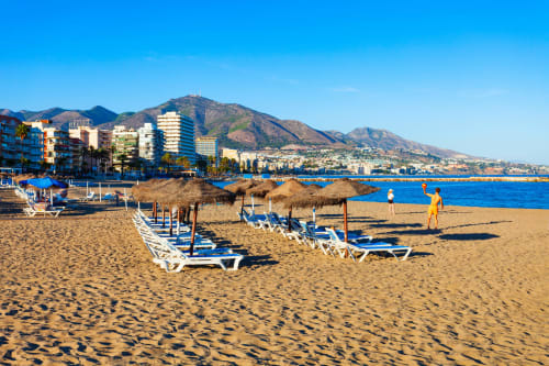 Playa de Fuengirola con hamacas azules, sombrillas de paja y sierra al fondo bajo cielo despejado