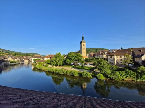 Village riverside d'Ornans avec clocher, collines verdoyantes et bâtiments historiques reflétés dans les eaux calmes de la rivière