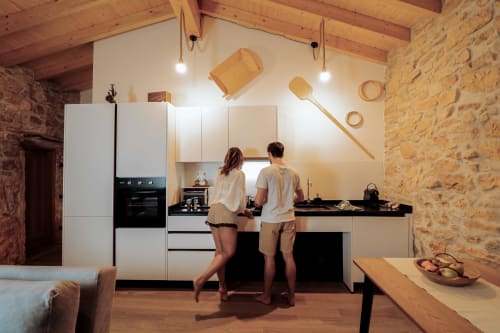 Casal na cozinha moderna de Recantos D'Almerinda, Pombal com armários brancos, paredes de pedra, vigas de madeira e luminárias pendentes