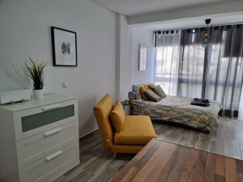 Dormitorio moderno en Fuengirola con silla mostaza, cama, cómoda blanca y puertas de terraza