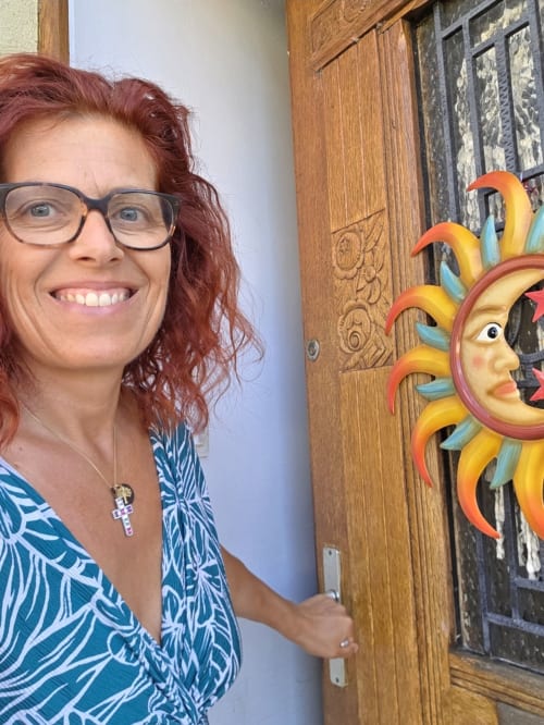 Smiling woman at Chez Mariebel' BnB in Ottonville, France, next to colorful sun mask decor
