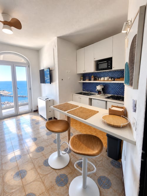 Cucina moderna con vista sul mare nella Divina Suite di Chiaia di Rose, Ischia. Arredamento bianco, rivestimento blu, sgabelli in legno.