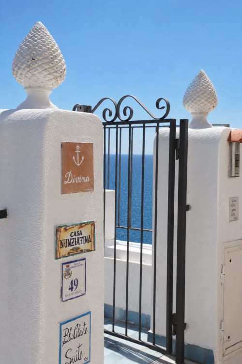 Ingresso di villa mediterranea imbiancata con decorazioni, cancello nero ornato e vista sul mare a Ischia, Italia