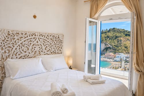 Camera da letto luminosa con testata scolpita, biancheria bianca e vista sul mare turchese a Chiaia di Rose, Ischia