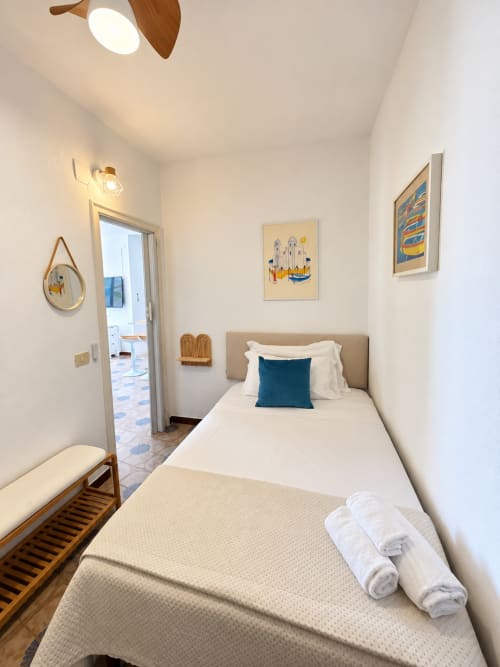 Camera luminosa nell'appartamento Chiaia di Rose a Ischia con letto singolo, cuscino turchese, panchina in legno e opere d'arte costiera