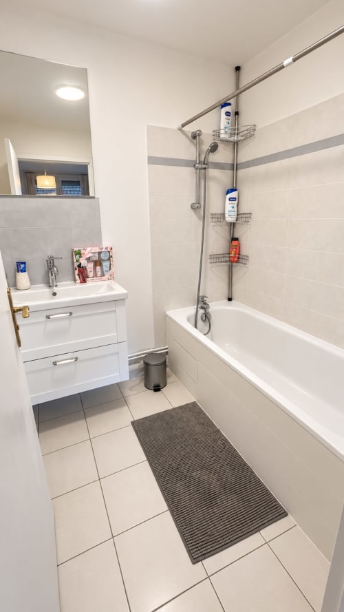 Salle de bain moderne avec vanité blanche, baignoire, douche avec rangements métalliques et carrelage beige chez Le Paradis Des Logis