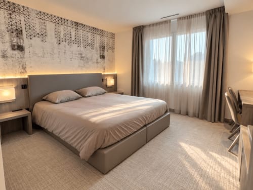 Chambre moderne aux Résidences Attica à St-Sulpice avec vue sur le lac, lit gris contemporain et voilages délicats.